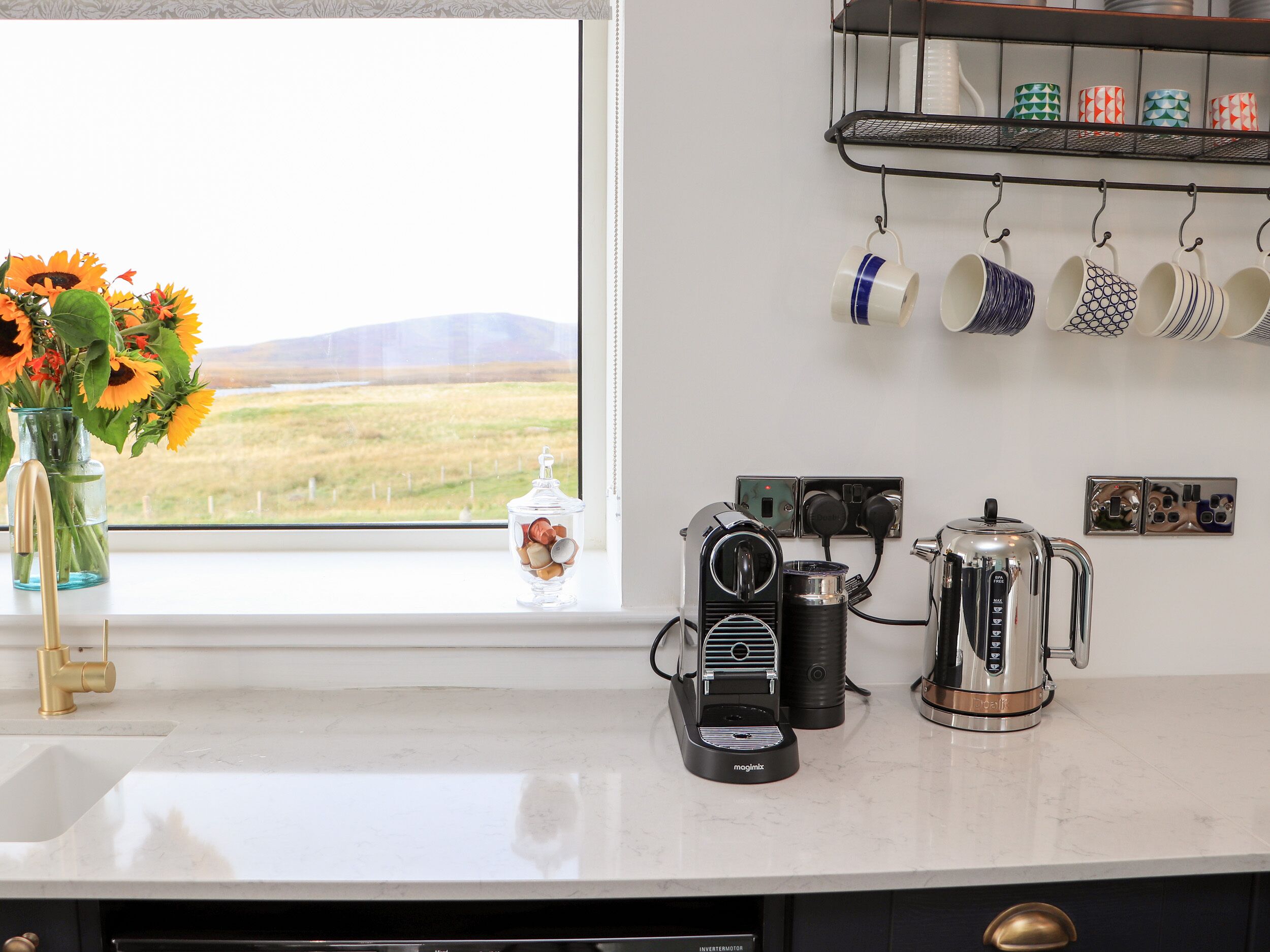 A kitchen with a window view, a coffee maker, a kettle, and a mug rack.