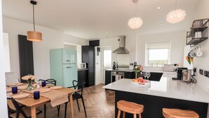 A modern kitchen with a central island, wooden stools, a dining table set with plates, and a microwave. - Park Cottage (Isle of North Uist)