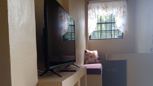 A living room with a flat-screen TV on a stand, a sofa, a window with floral curtains, and a tiled floor. - Near Ferry, Airport, Shops, Beaches, Atms, Rodney Bay and Castries - Free Pickup (Castries)