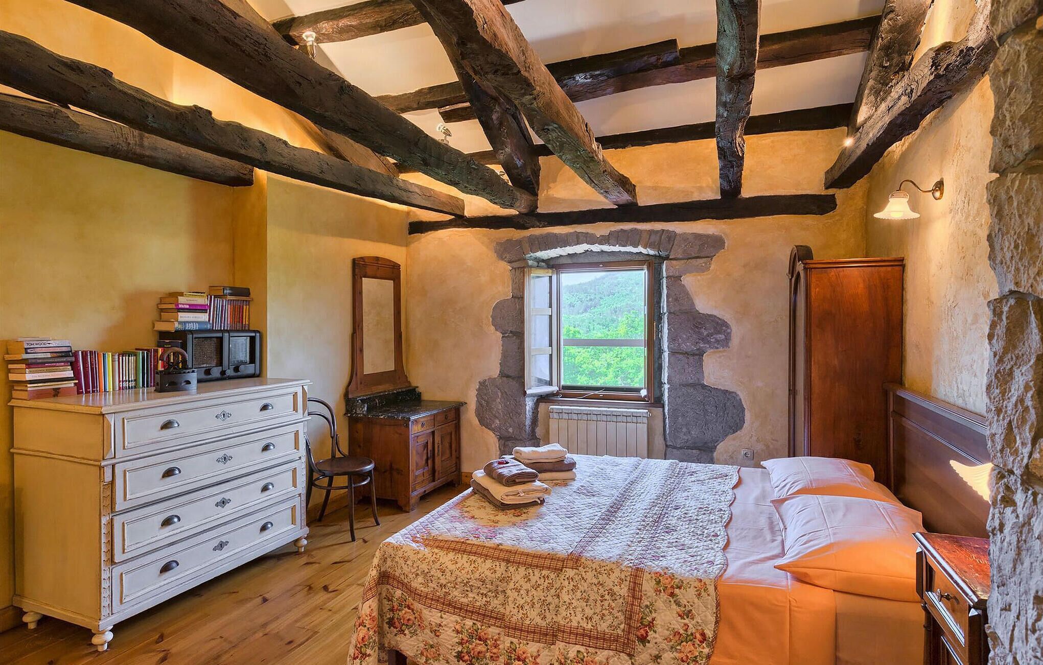 A rustic bedroom with exposed wooden beams, a stone wall, a window with a view of greenery, a dresser, a chair, and a bed with a quilt.