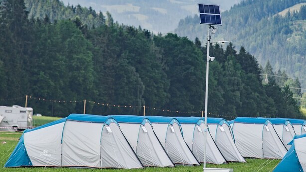 A row of tents in a grassy field with a solar panel and a signpost in the foreground, set against a backdrop of mountains and a partly cloudy sky.