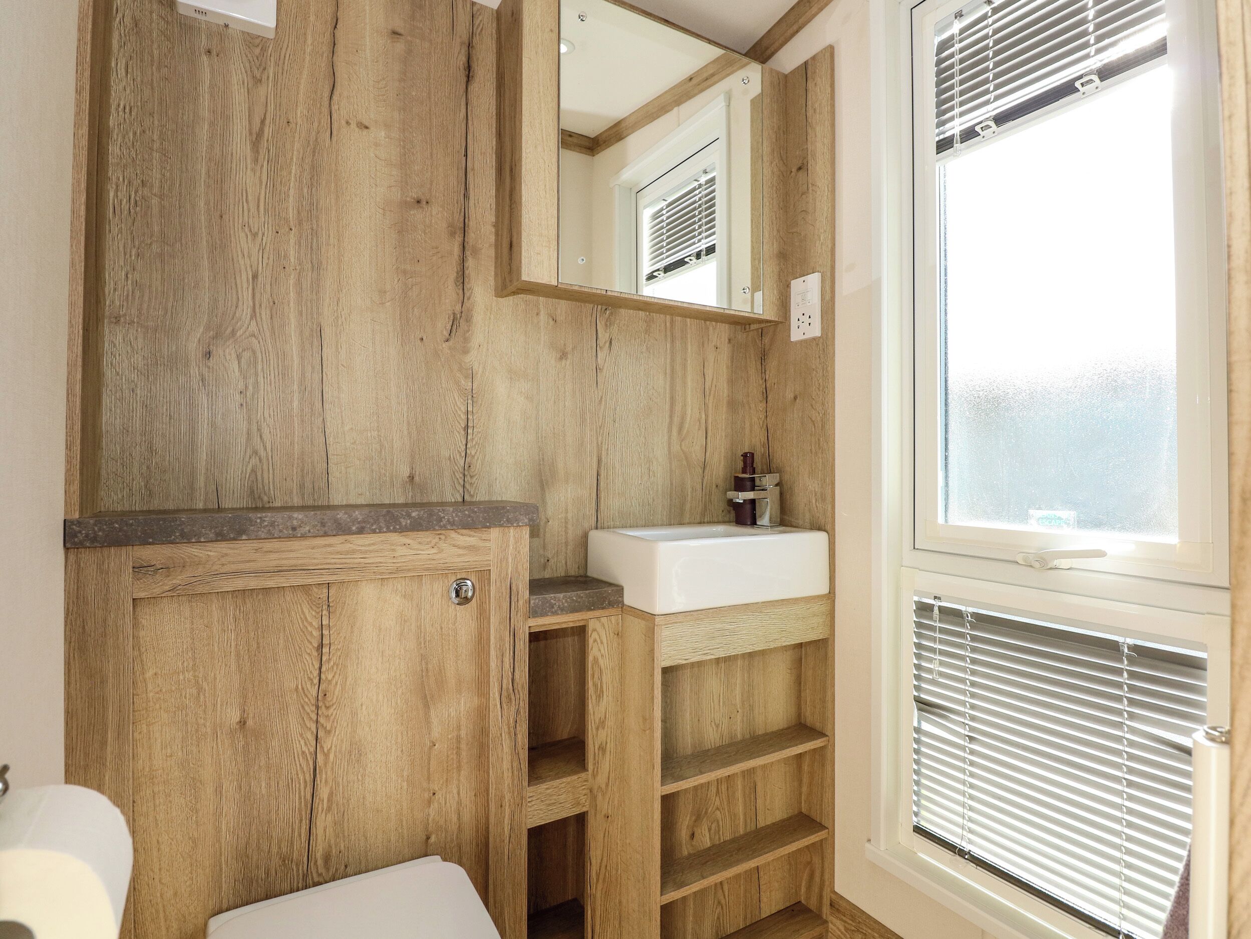 Une salle de bain avec des murs en bois, un lavabo blanc et une fenêtre avec des stores.