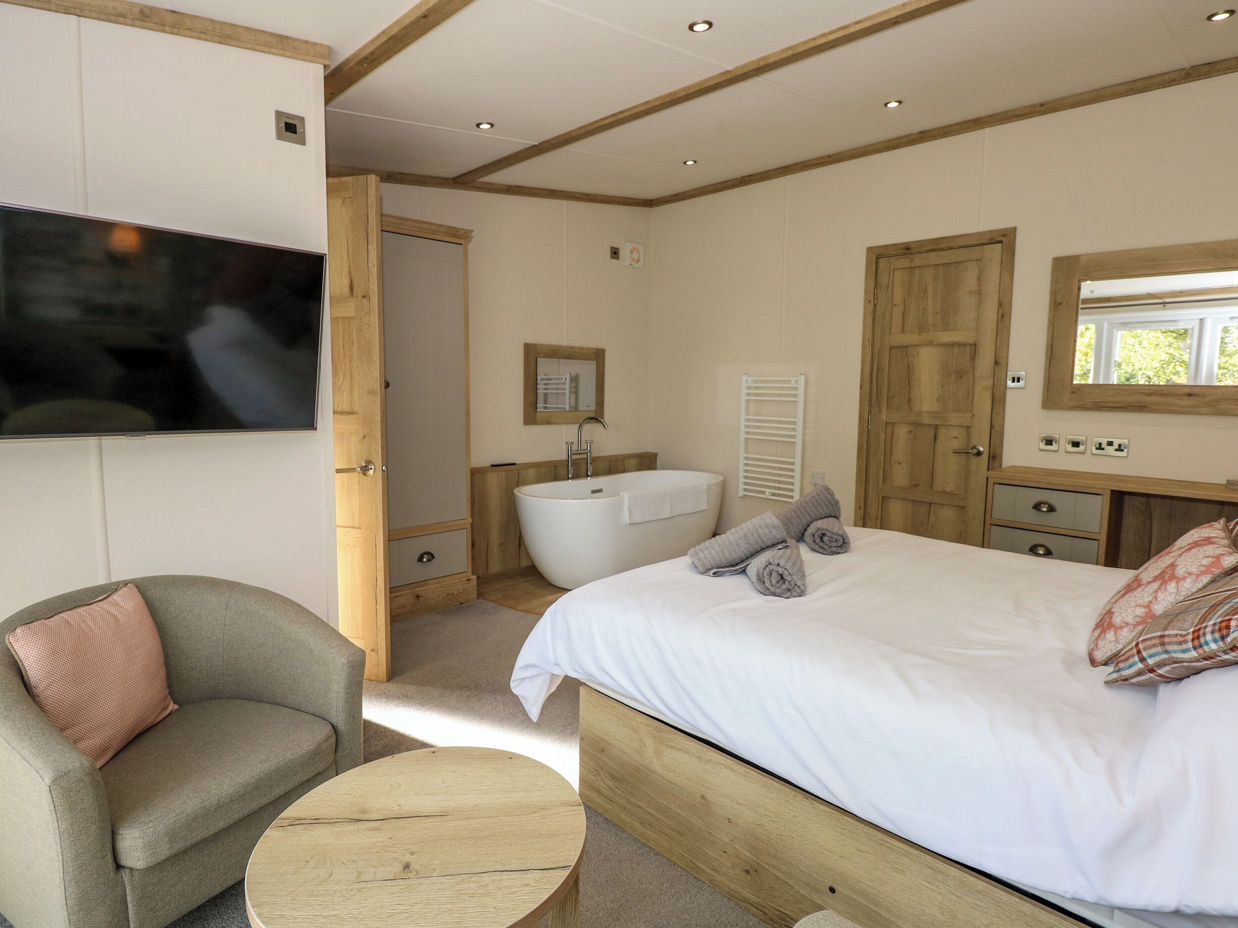 Un dormitorio con cama, una silla, una mesa redonda de madera, un televisor montado en la pared, un baño con bañera y un armario de madera.