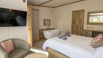 Un dormitorio con cama, una silla, una mesa redonda de madera, un televisor montado en la pared, un baño con bañera y un armario de madera.