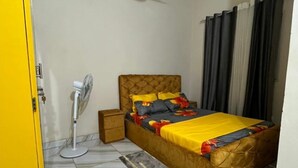 A bedroom with a bed, a standing fan, a nightstand, a yellow door, and a ceiling fan. - Residence in Tounkaranké (Bamako)