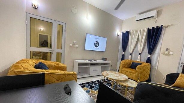 A modern living room with two yellow sofas, a coffee table, a TV mounted on the wall, and a ceiling fan. - Residence in Tounkaranké (Bamako)