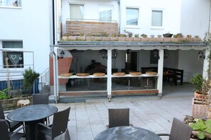 An outdoor patio area with round tables and wicker chairs, a covered seating area with cushions, and a brick wall with potted plants. - Rustic Apartment in Essen (Essen)