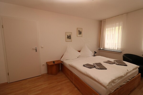 Un dormitorio con cama de madera, almohadas blancas y una mesita de noche con control remoto. Hay dos cuadros en la pared y una ventana con cortinas de encaje.