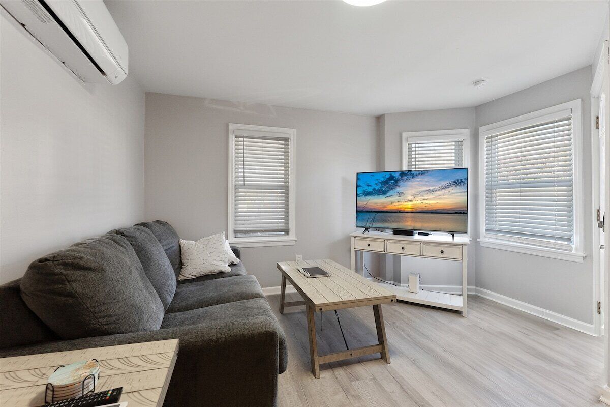 A living room with a grey sofa, a wooden coffee table, a flat-screen TV on a white stand, and a painting on the wall.