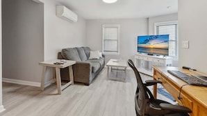 A modern living room with a grey sofa, a small table, a TV, and a wooden desk with a chair. - Modern 2BR Condo Walk to Beach and Dining (Wildwood)