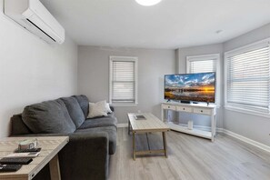 Uma sala de estar moderna com um sofá cinzento, uma mesa de centro de madeira, uma televisão de tela plana e um rack branco para eletrônicos.