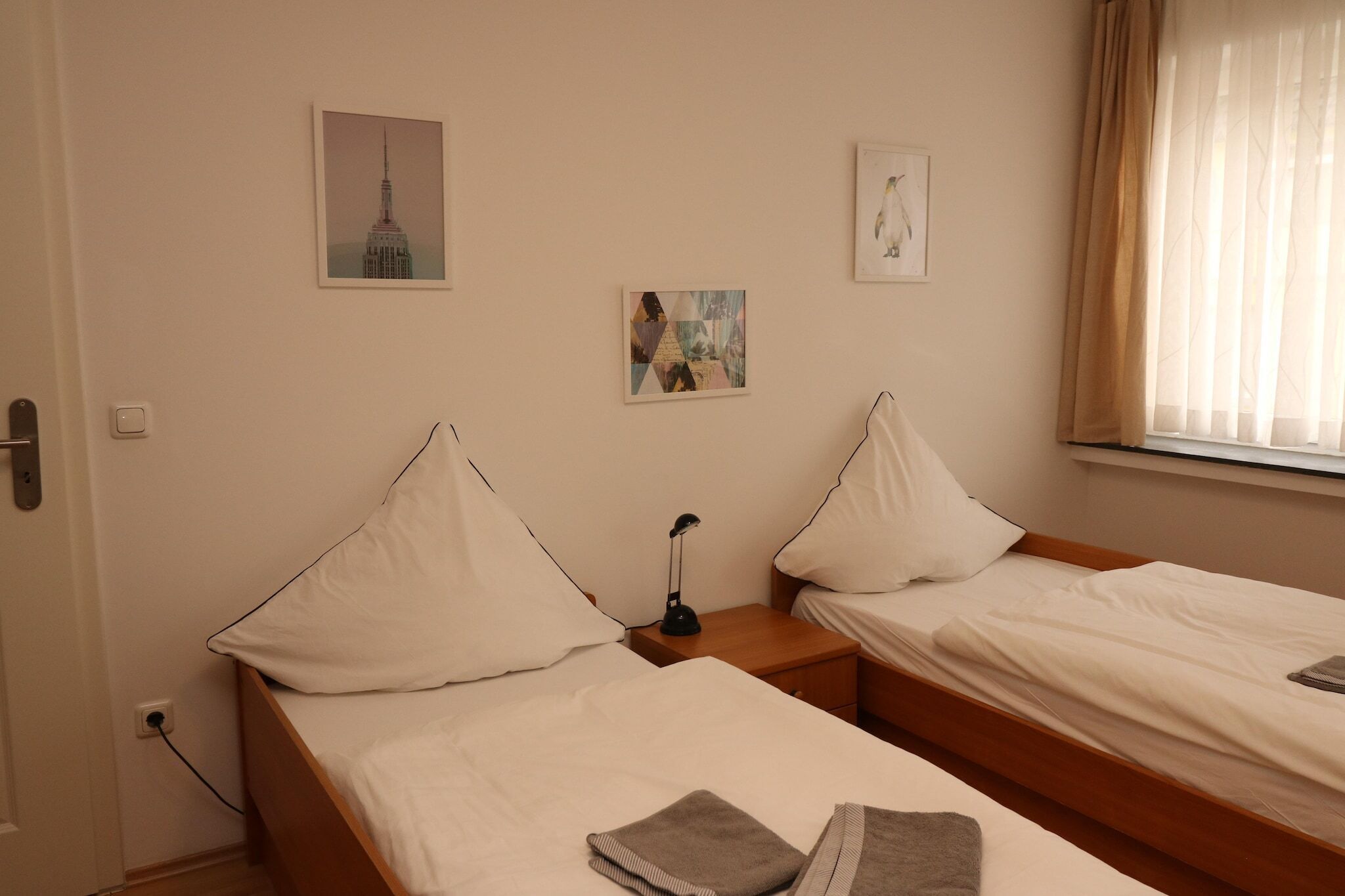 A hotel room with two single beds, a nightstand with a lamp, and artwork on the walls.