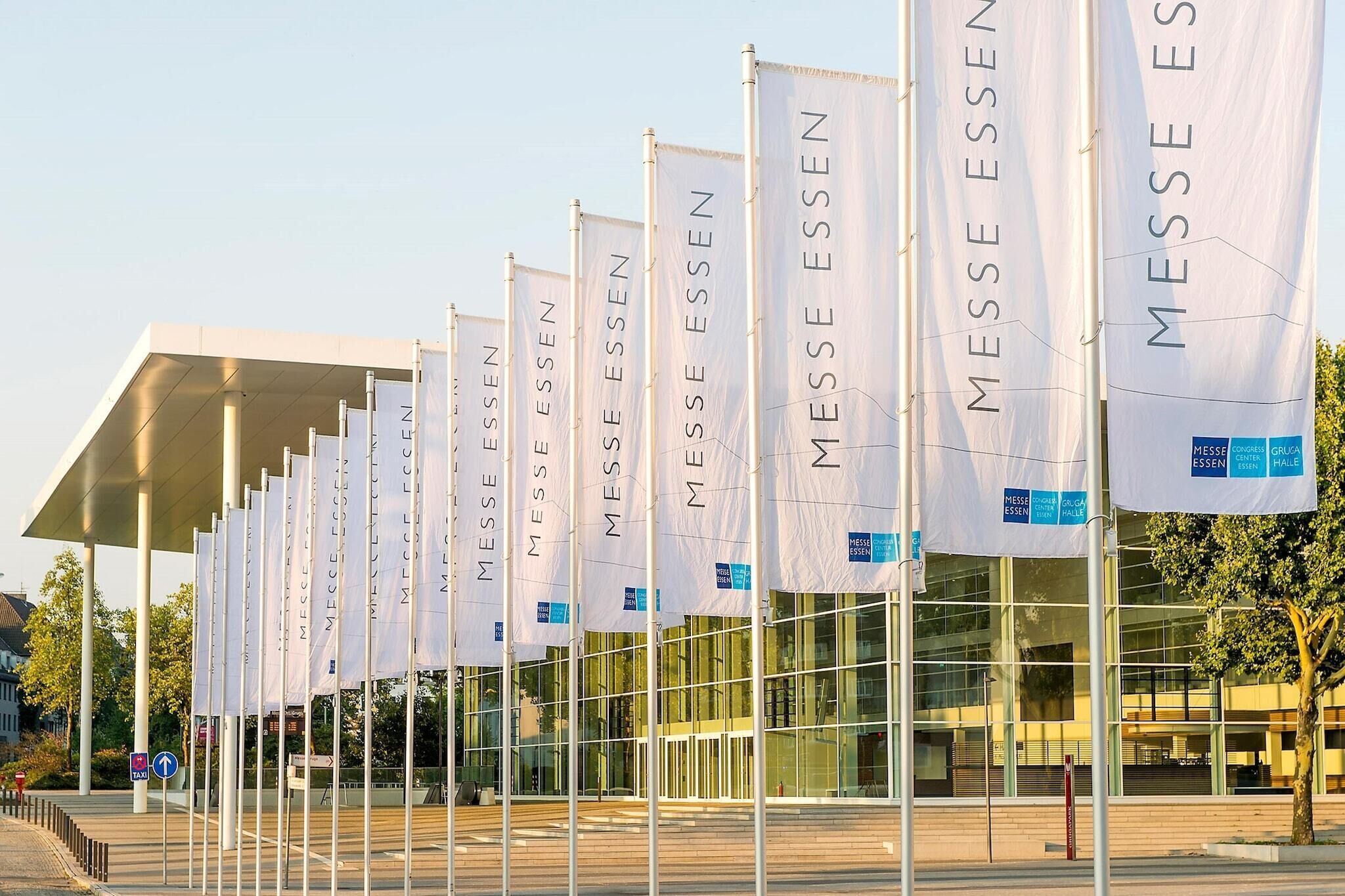 Un edificio moderno con varias banderas blancas que muestran 'MESSE ESSEN' y 'HALL MESSE ESSEN'.
