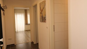 Un couloir avec une porte donnant sur une pièce dotée d’une grande fenêtre et de rideaux. Un tableau est accroché au mur et un porte-manteau se trouve sur la gauche.