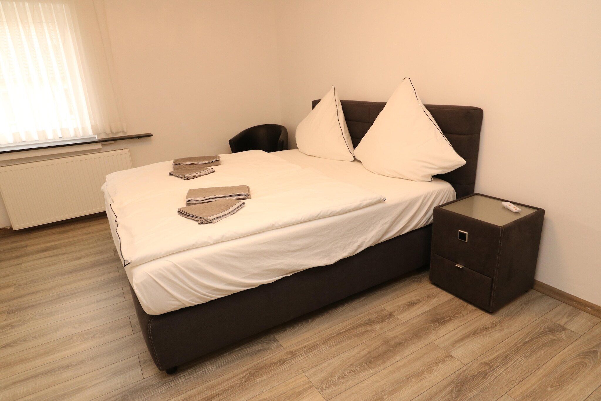 A neatly made bed with a dark headboard and two pillows, a small bedside table with a remote control, and a radiator in the background.
