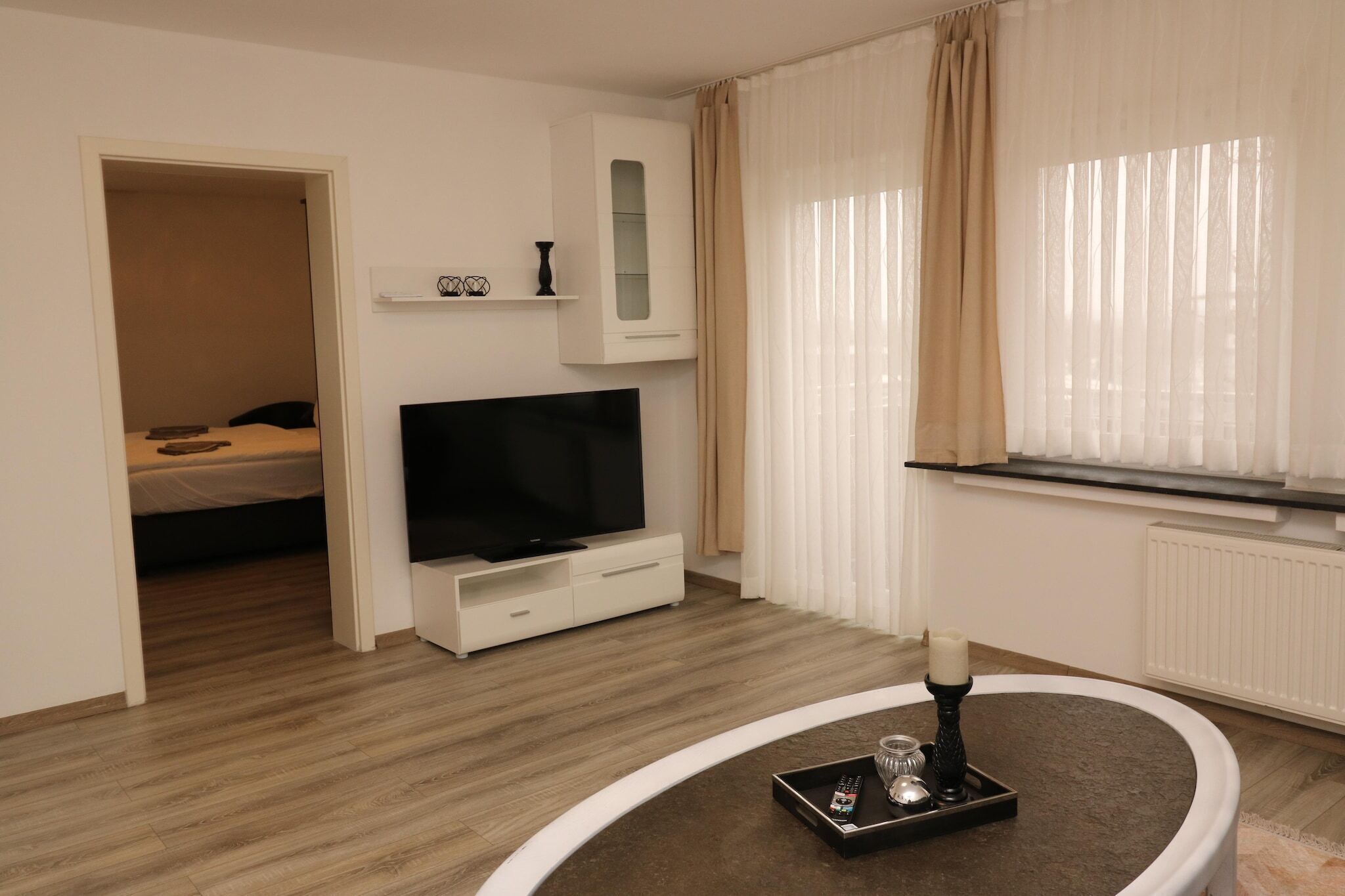 Une chambre d’hôtel moderne équipée d’un téléviseur à écran plat, d’une petite armoire, d’une table de chevet avec un plateau, et d’une fenêtre avec des rideaux légers.