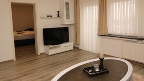 Une chambre d’hôtel moderne équipée d’un téléviseur à écran plat, d’une petite armoire, d’une table de chevet avec un plateau, et d’une fenêtre avec des rideaux légers.
