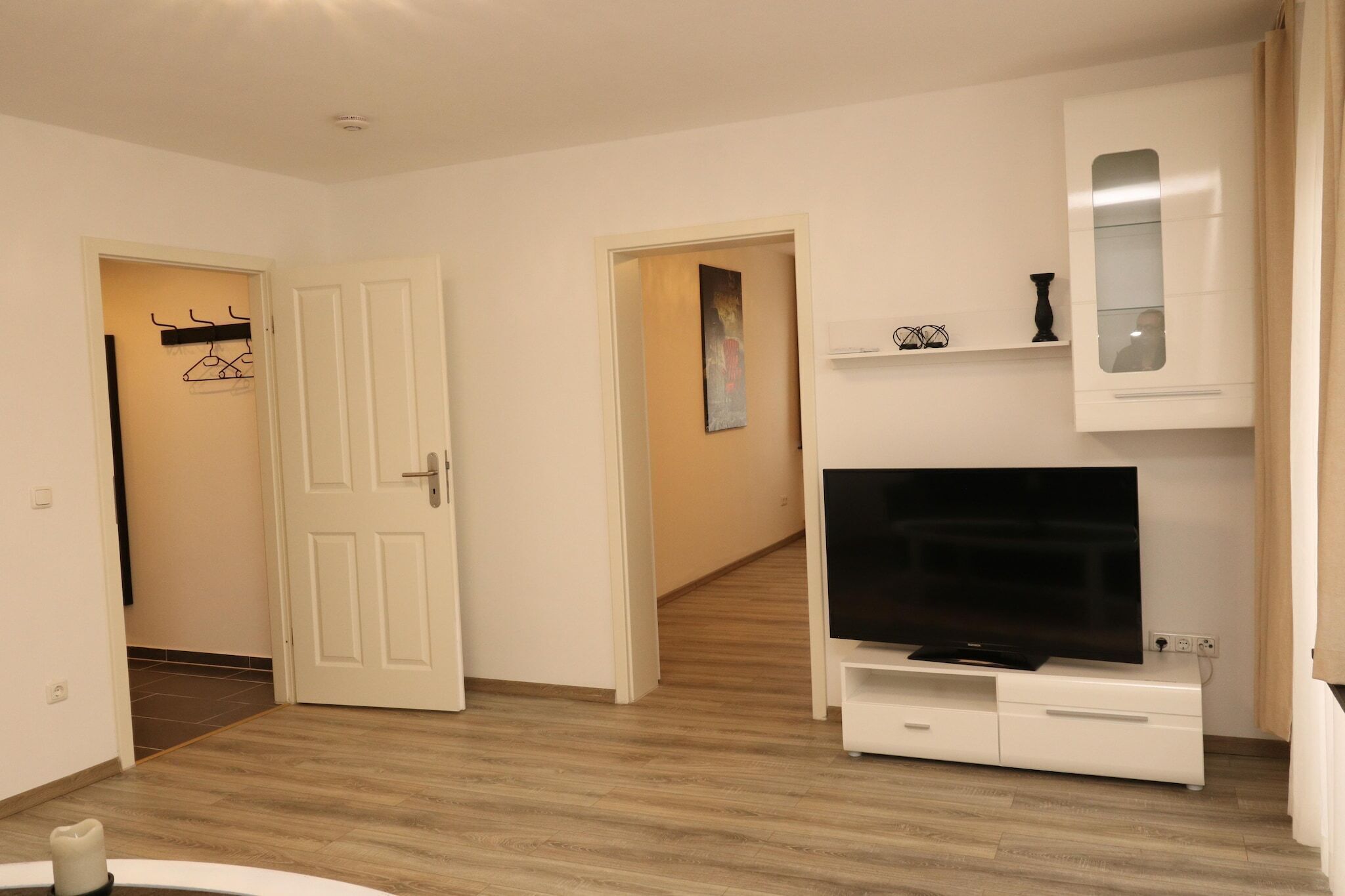 Un salon moderne avec un téléviseur à écran plat posé sur un meuble blanc, un couloir menant à une autre pièce et une porte avec un porte-manteau.