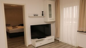 Une chambre d’hôtel moderne avec un grand téléviseur à écran plat posé sur un meuble blanc, un lit recouvert d’une courtepointe beige et une petite étagère ornée d’objets décoratifs.