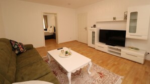 Ein Wohnzimmer mit einem grünen Sofa, einem weißen Couchtisch, einem Flachbildfernseher auf einer weißen Unterhaltungseinheit und einer Tür zu einem anderen Raum.
