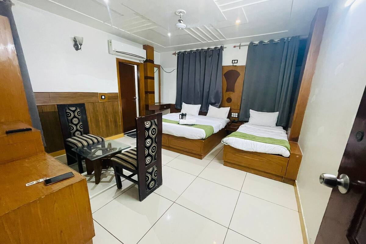 Une chambre d’hôtel avec deux lits en bois, une petite table avec des chaises, un miroir et un ventilateur de plafond.