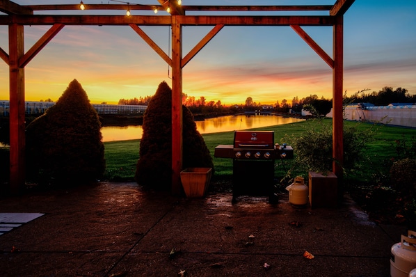 A covered outdoor area with a grill and propane tanks, overlooking a lake and trees during sunset.