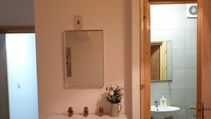 Quarto com cama, penteadeira, espelho, uma pequena planta e banheiro visível por uma porta aberta.