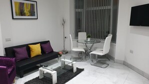 Uma sala de estar moderna com um sofá preto, cadeiras brancas, uma mesa de centro de vidro e uma janela grande com persianas.