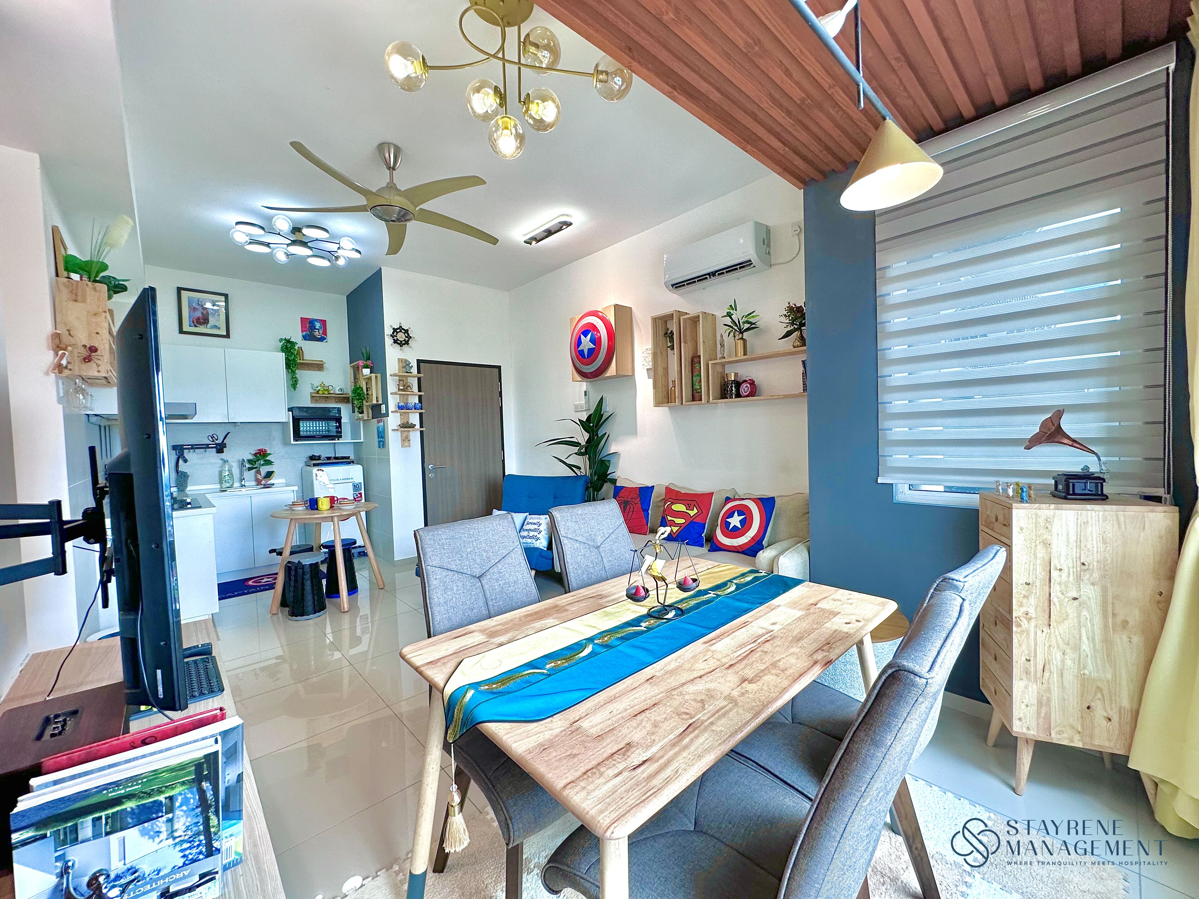 A modern living room with a wooden dining table, a ceiling fan, and a sofa with superhero-themed cushions.