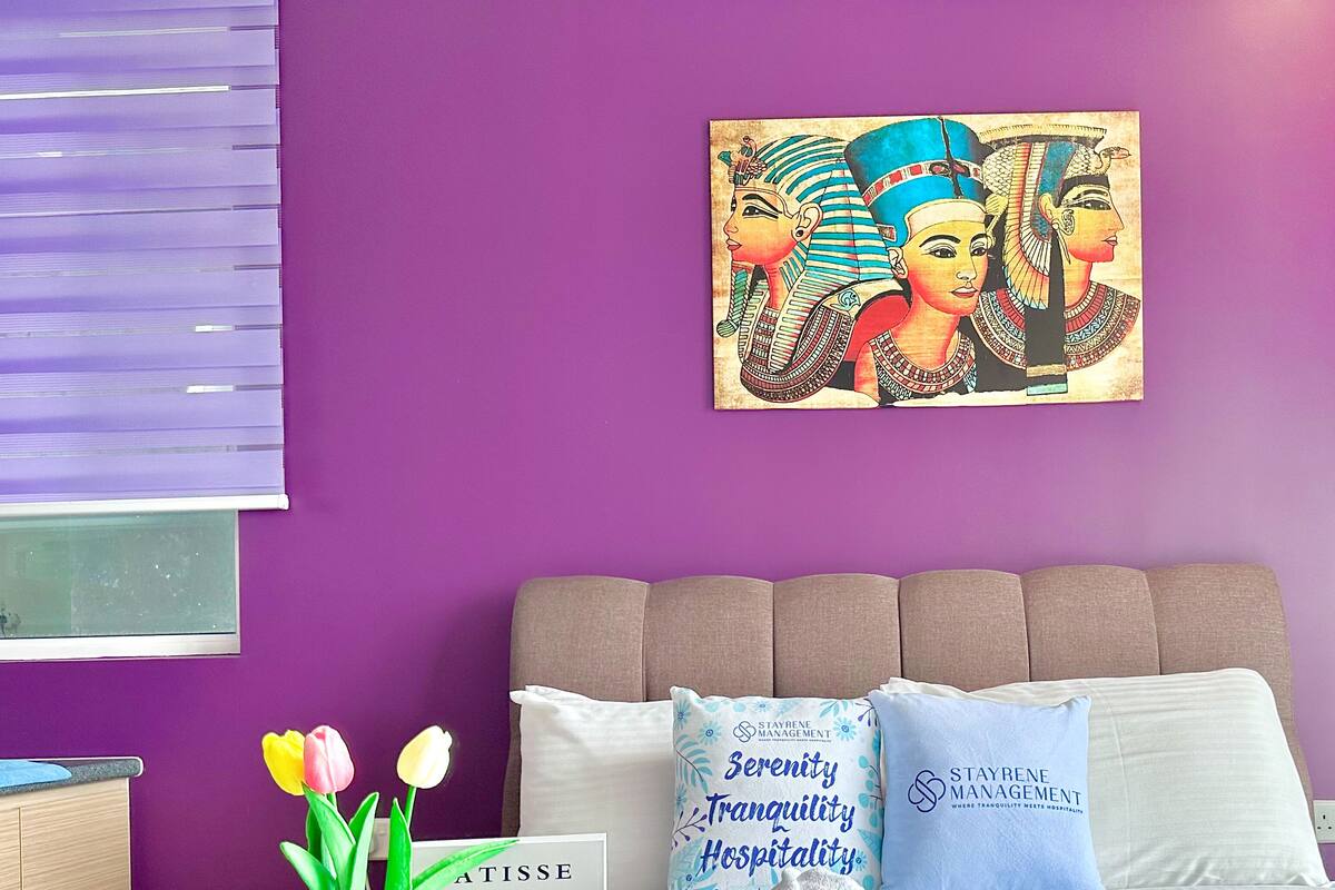 Ein Hotelzimmer mit lila Wand, einem Bett mit weißen und blauen Kissen, einem Gemälde, einer Vase mit Tulpen und einem Tablett mit Handtuch und einem gerahmten Bild.