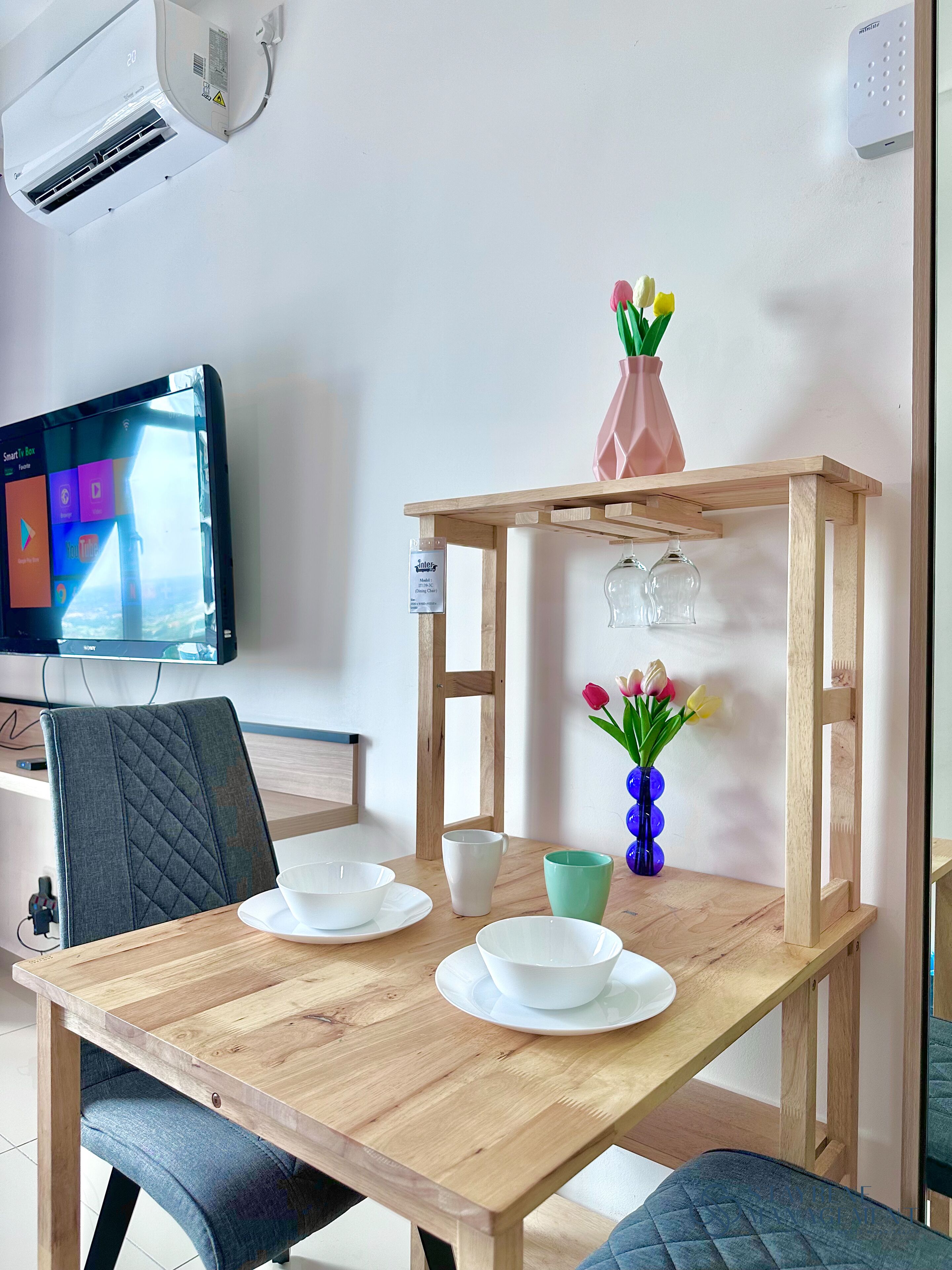 A wooden dining table with white bowls and a green cup, a vase with flowers, and a mounted television.