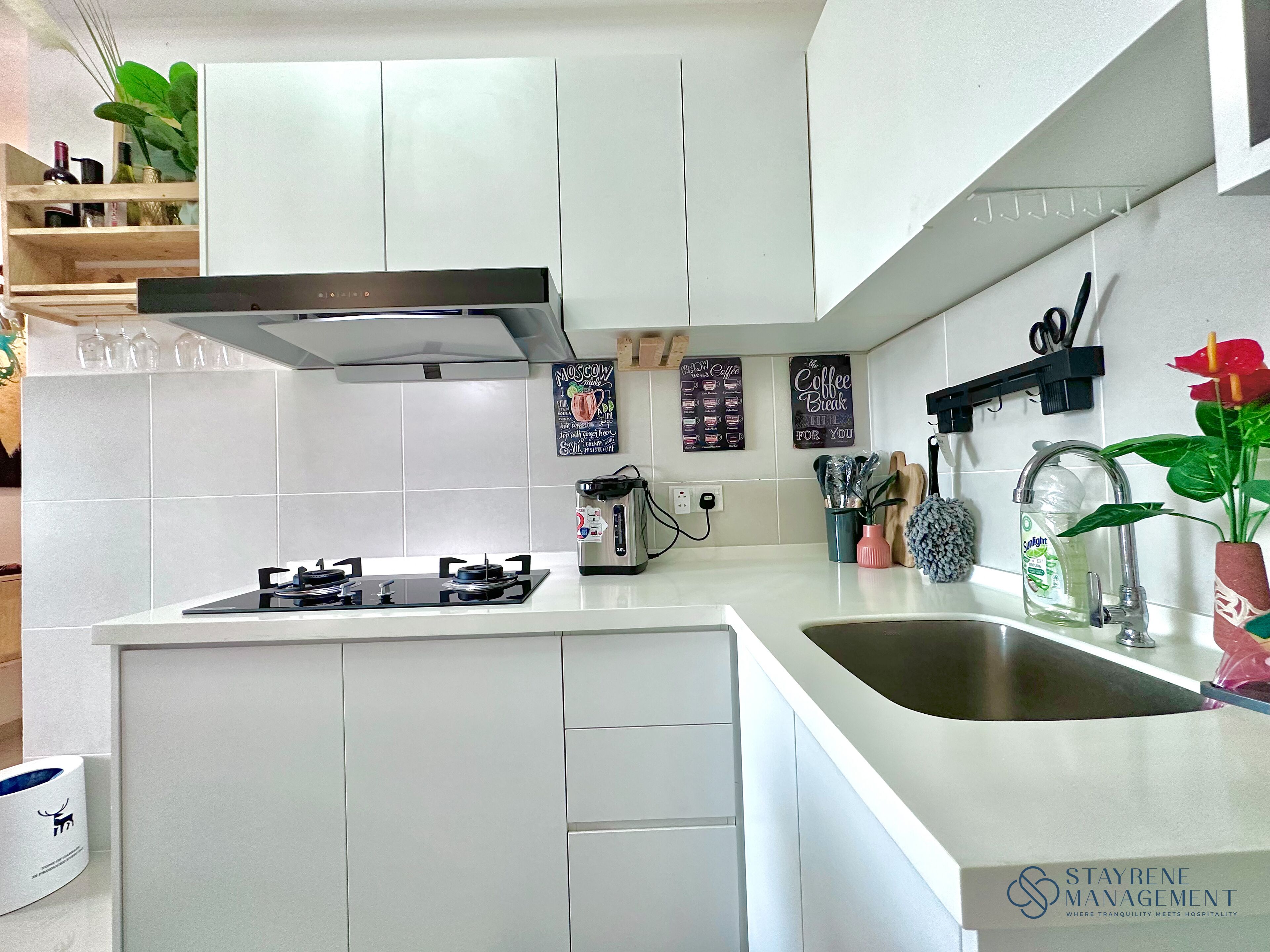 A modern kitchen with white cabinetry, a gas stove, a coffee machine, and a sink. There are decorative items like a plant and a sign that says "Coffee Break".
