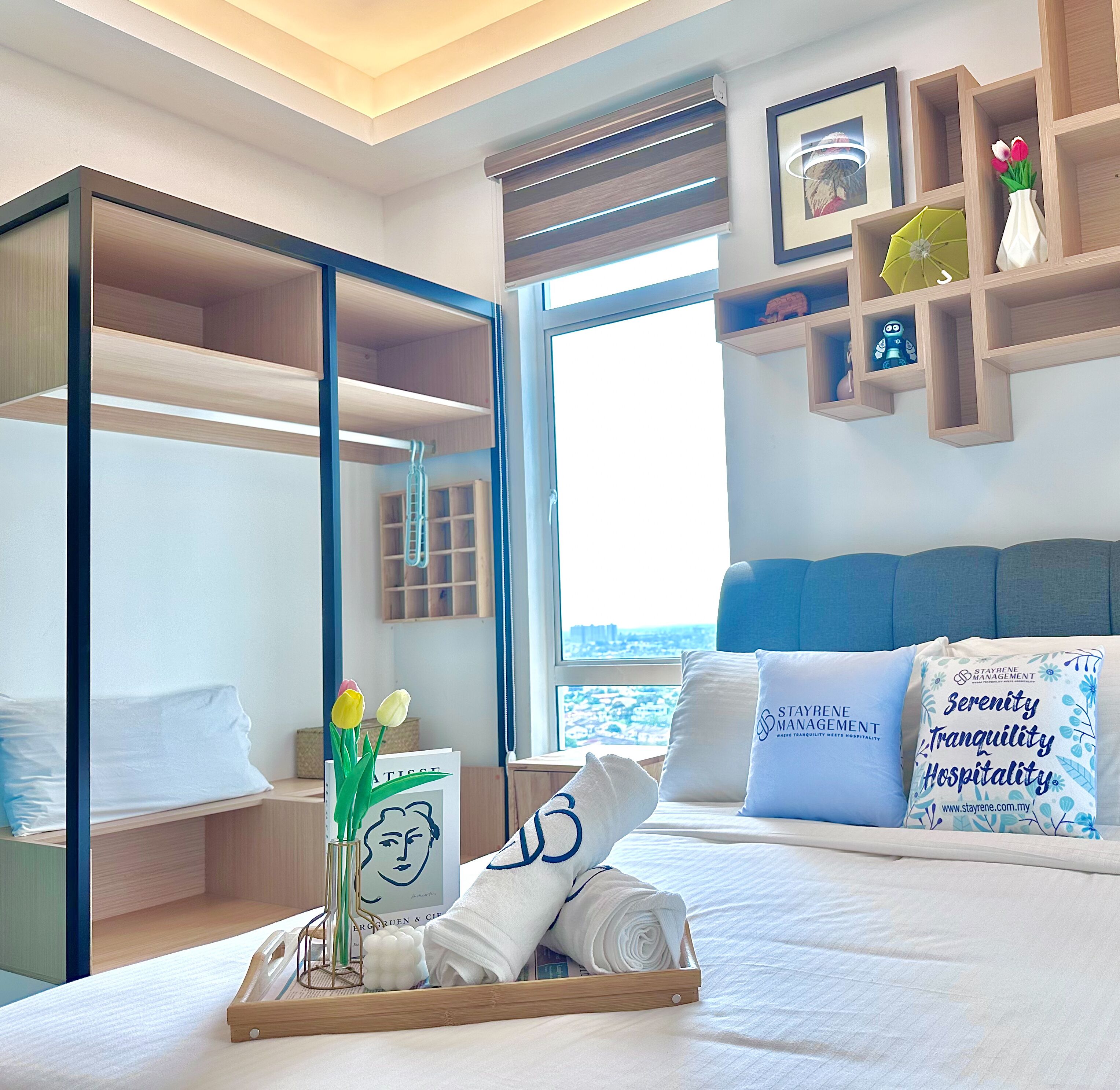 A hotel room with a bed, a desk, a window with a city view, a shelf with decorative items, and a framed picture on the wall.