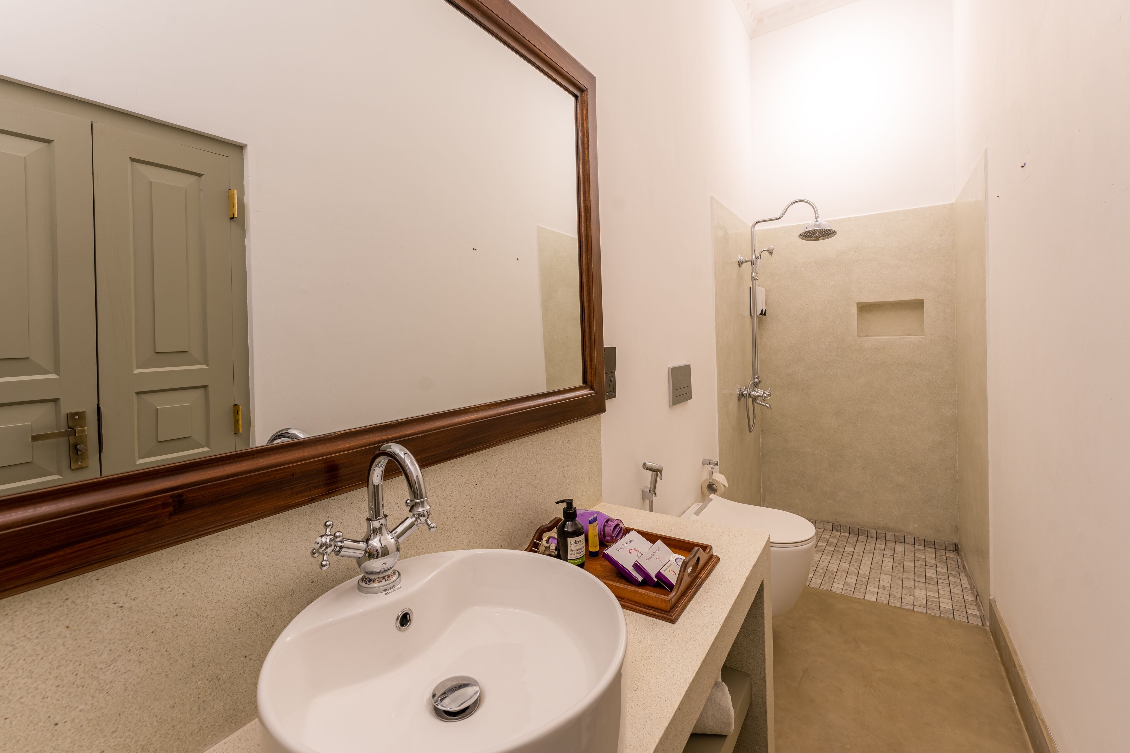 A bathroom with a white sink, a mirror, a shower, and a wooden tray with toiletries.