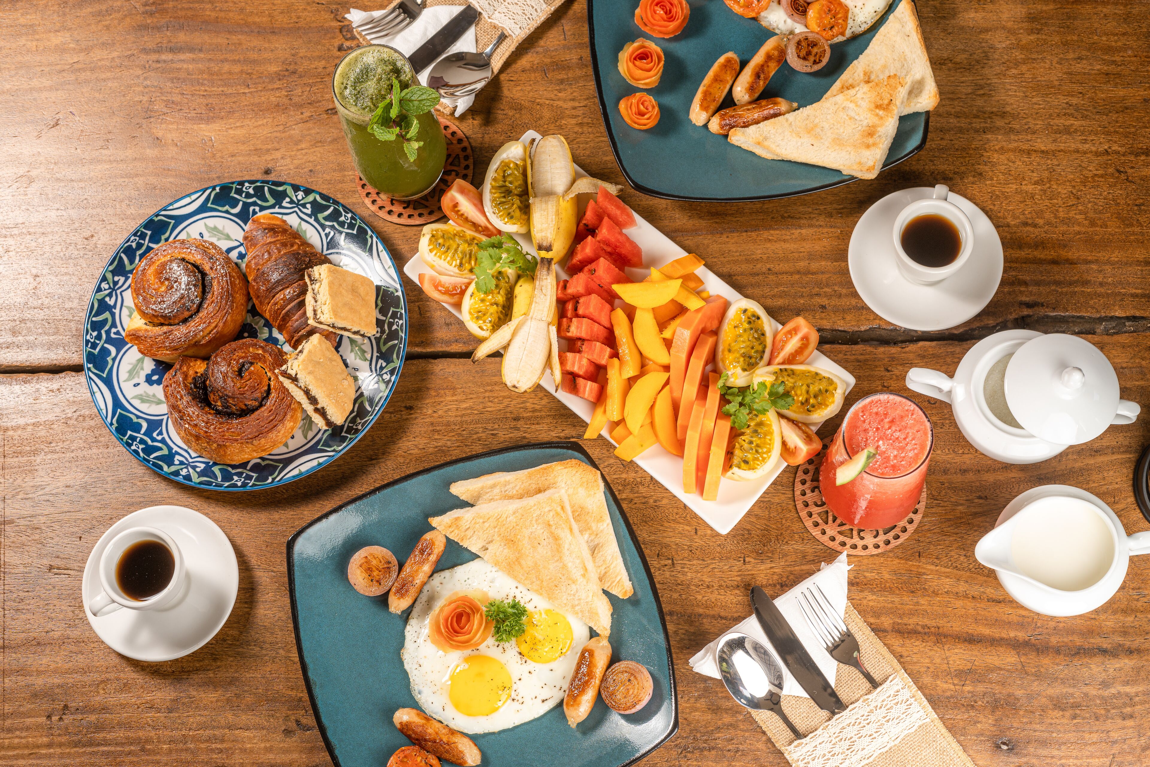 A breakfast spread with croissants, eggs, sausages, and various fruits and vegetables on a wooden table.
