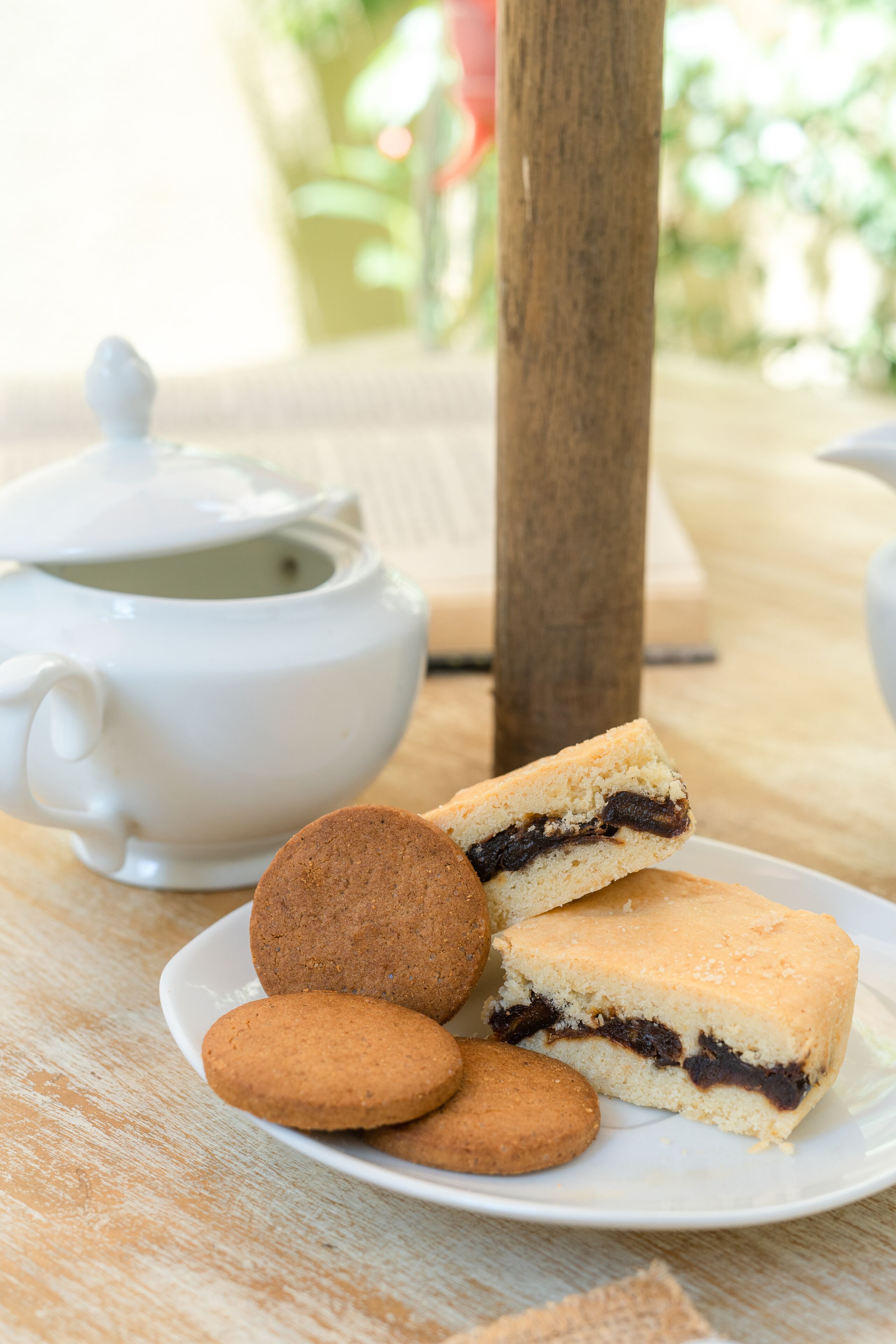 A white teapot and a plate with cookies and a sandwich on a wooden table.