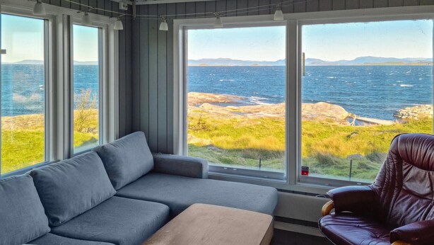 A room with a grey sofa, a brown ottoman, and a leather chair, with large windows offering a view of the sea and rocky landscape. - Amazing home in Rennesøy with house sea view (Rennesøy)