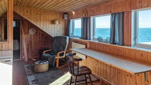 A cabin with wooden walls and a large window overlooking the sea. There is a wooden bench, a chair, and a small table. - Amazing home in Rennesøy with house sea view (Rennesøy)
