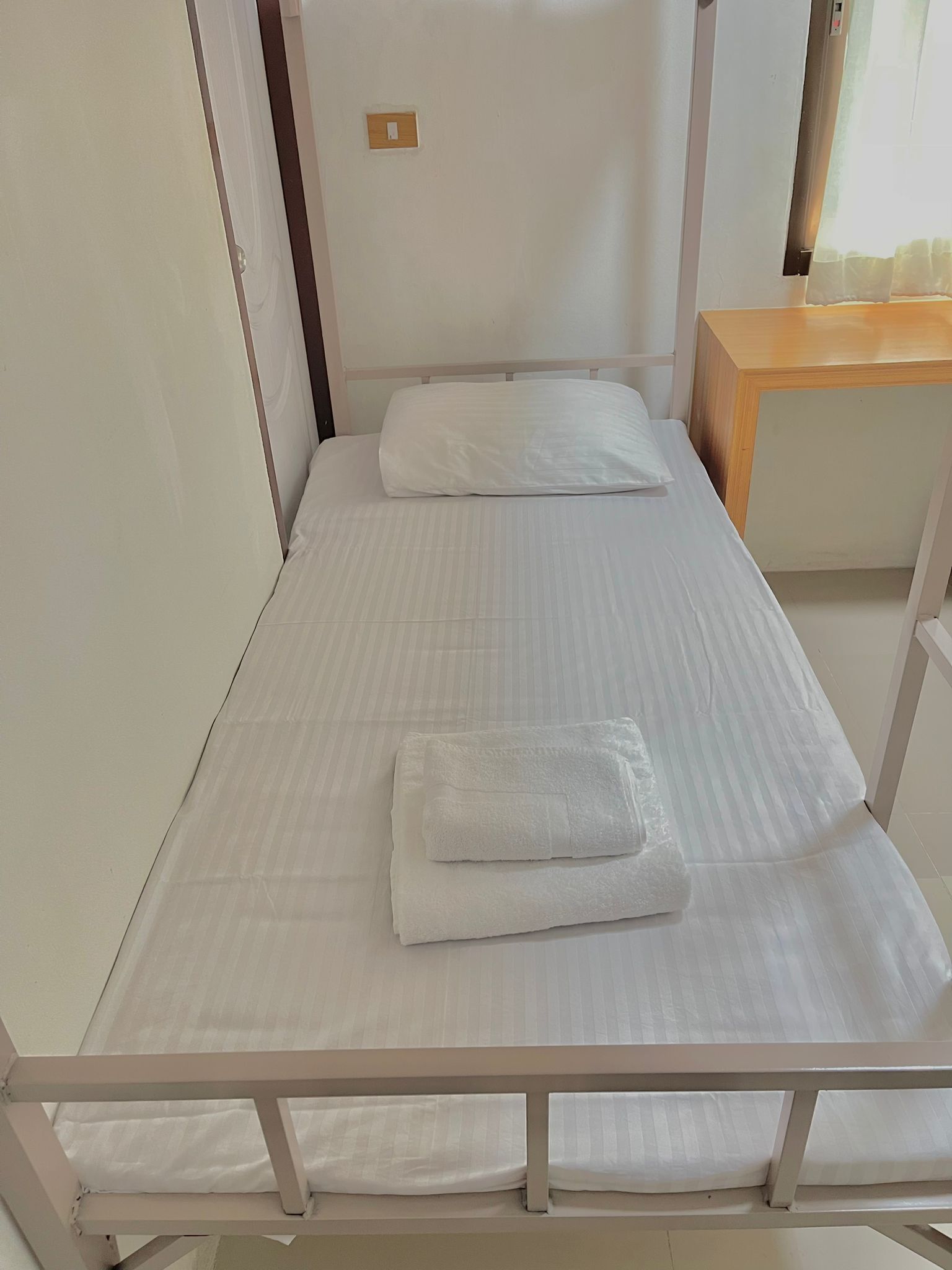 Una cama individual con ropa de cama blanca, una mesita de noche de madera y un interruptor de luz pequeño en la pared.