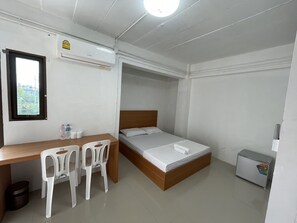 A small, clean room with a wooden desk, two white plastic chairs, a bed with a wooden headboard, a small refrigerator, and an air conditioning unit on the wall. - OYO 75577 Palace Hostel (Bangkok)