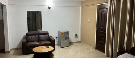 A room with a bed, a brown leather sofa, a small table, a refrigerator, and a door with curtains.