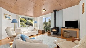 Ein gemütliches Wohnzimmer mit einer Couch, Sesseln, einem Fernseher, einem Kamin und großen Fenstern mit Blick auf Grünflächen.