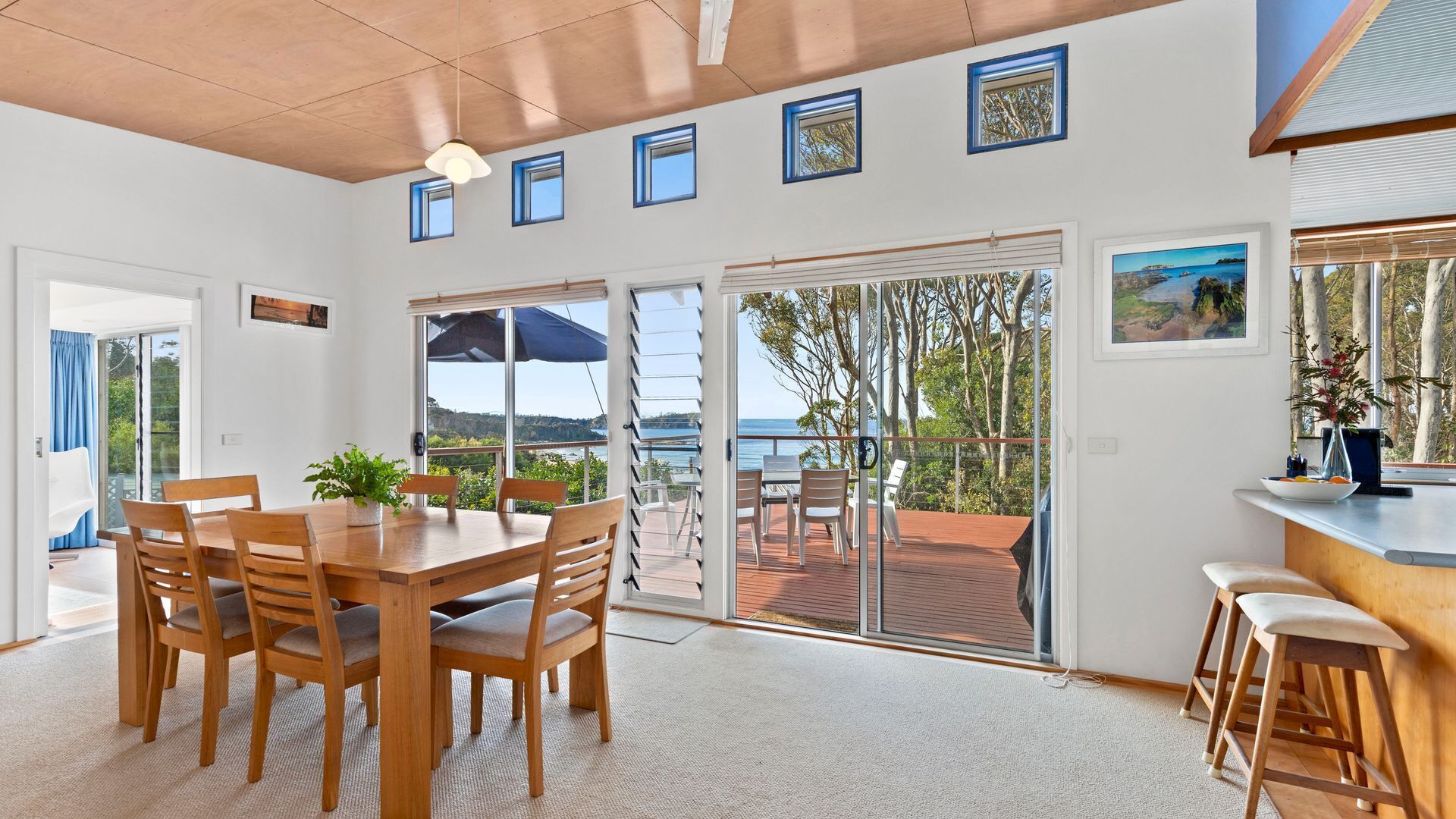 A spacious room with a large sliding glass door leading to an outdoor deck with a view of the sea. There is a wooden dining table with chairs and a small bar area with stools.