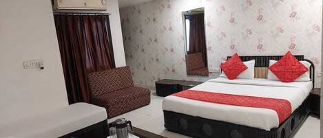 A hotel room with a bed, a sofa, a small table, an air conditioning unit, a ceiling fan, and a mirror.