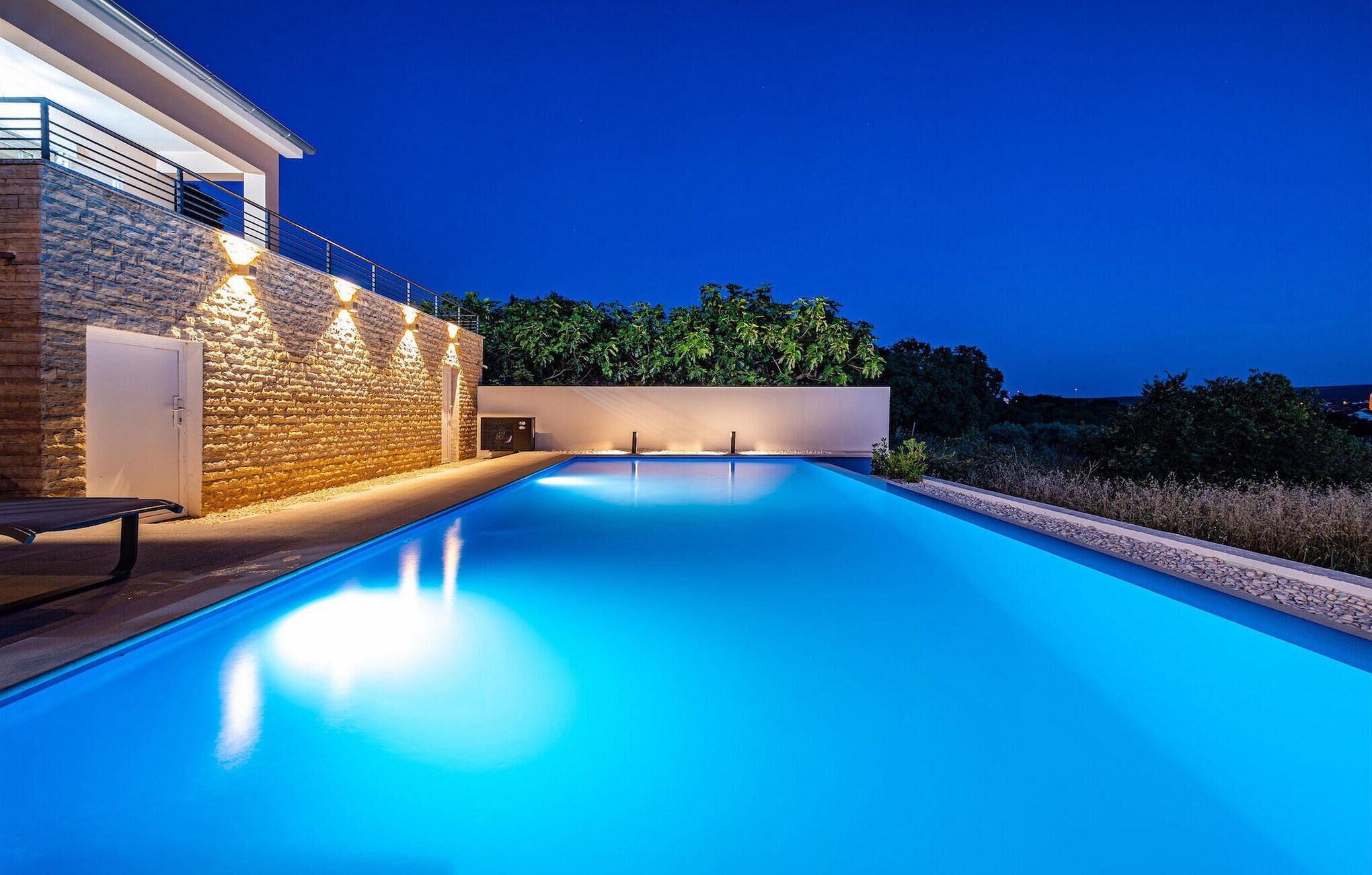 A modern pool area with a stone wall and a bench, surrounded by greenery.