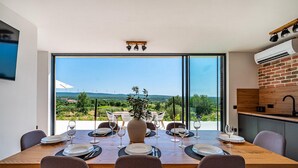 Un comedor con una gran mesa dispuesta para seis personas, con vistas a un paisaje pintoresco con colinas y aerogeneradores.
