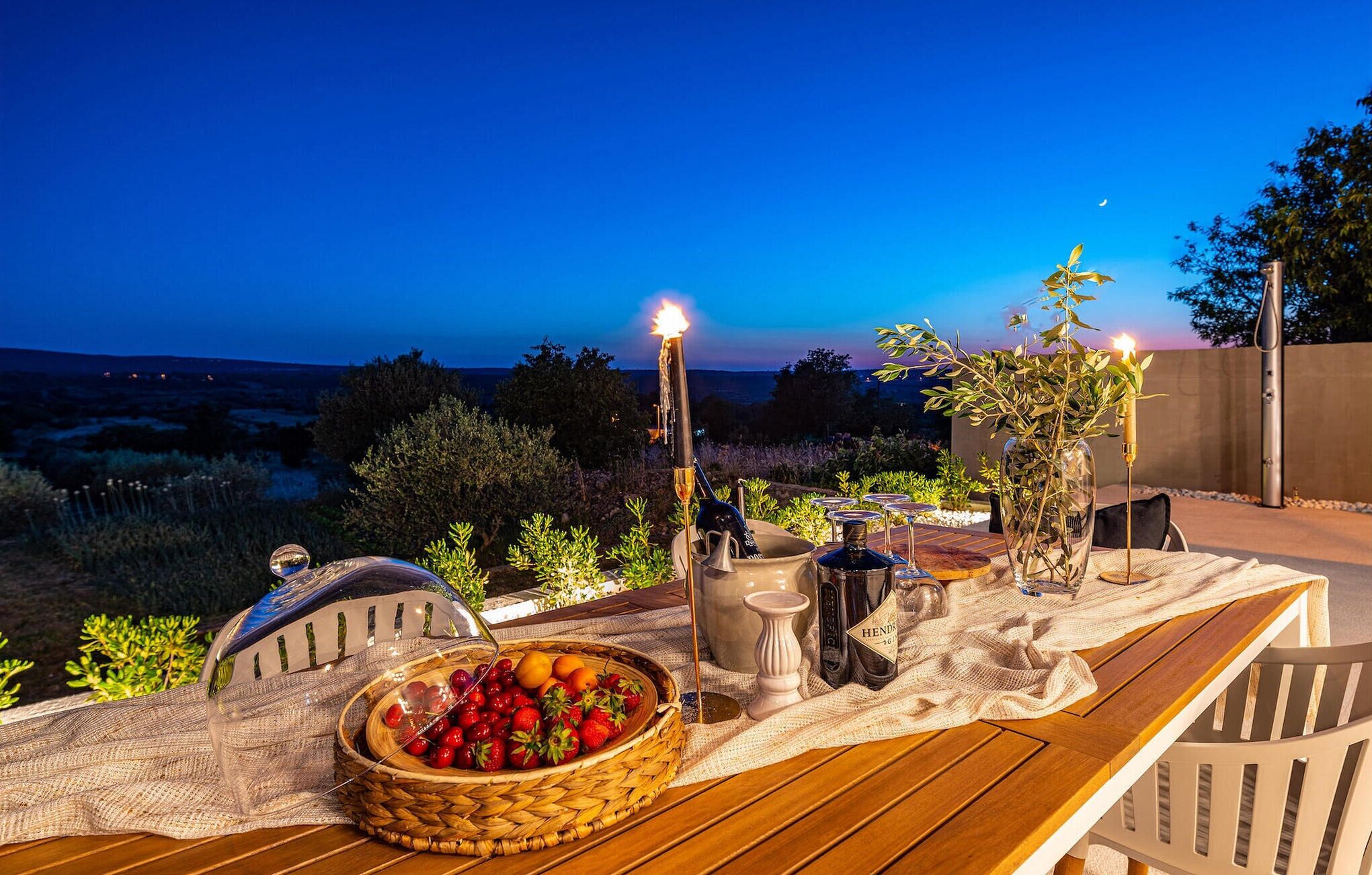 Un espace de restauration en plein air avec une table en bois, un panier de fruits, des bougies et une vue sur le paysage.