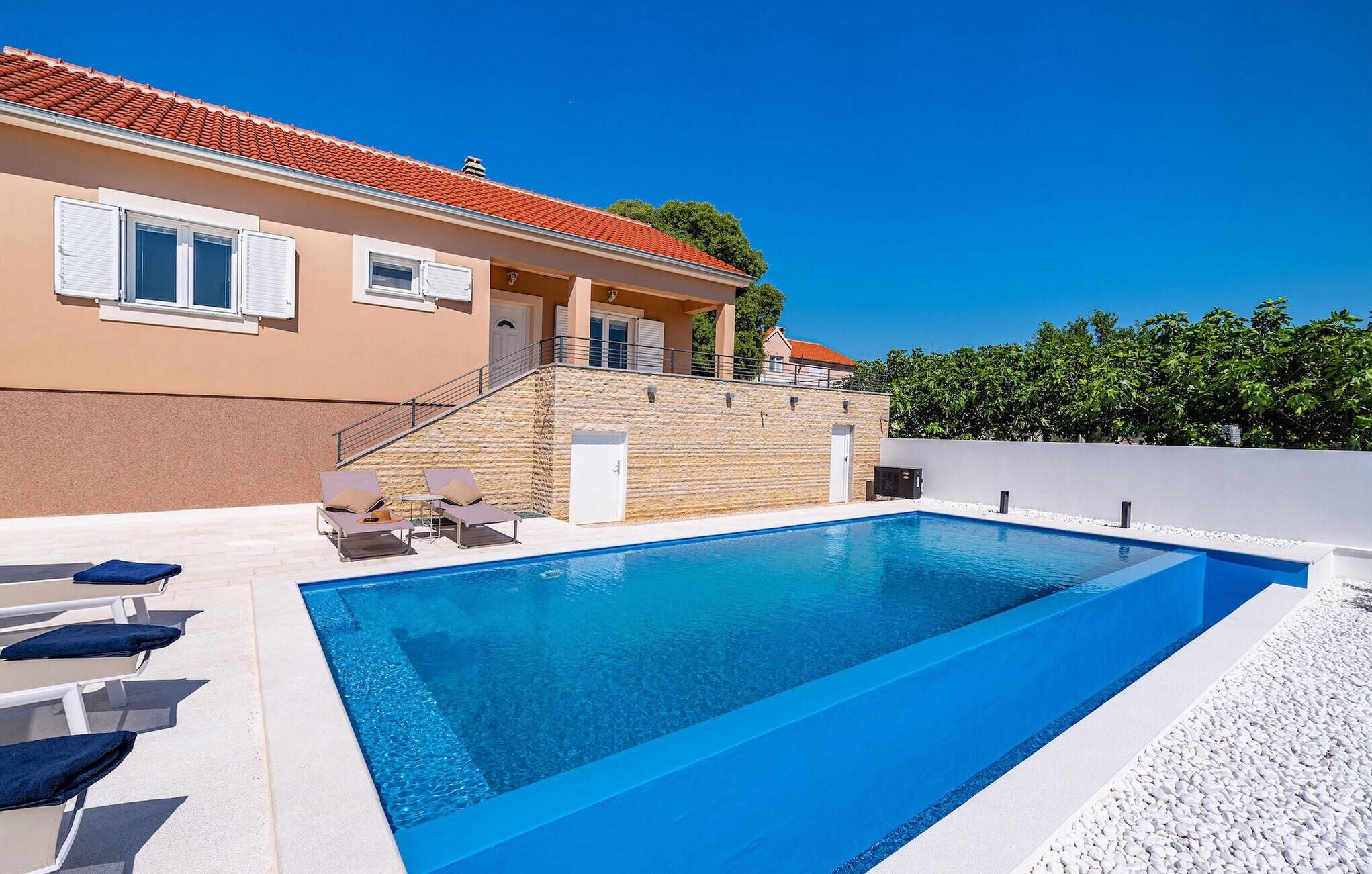 A swimming pool with lounge chairs, a stone wall, and a building with a red-tiled roof.