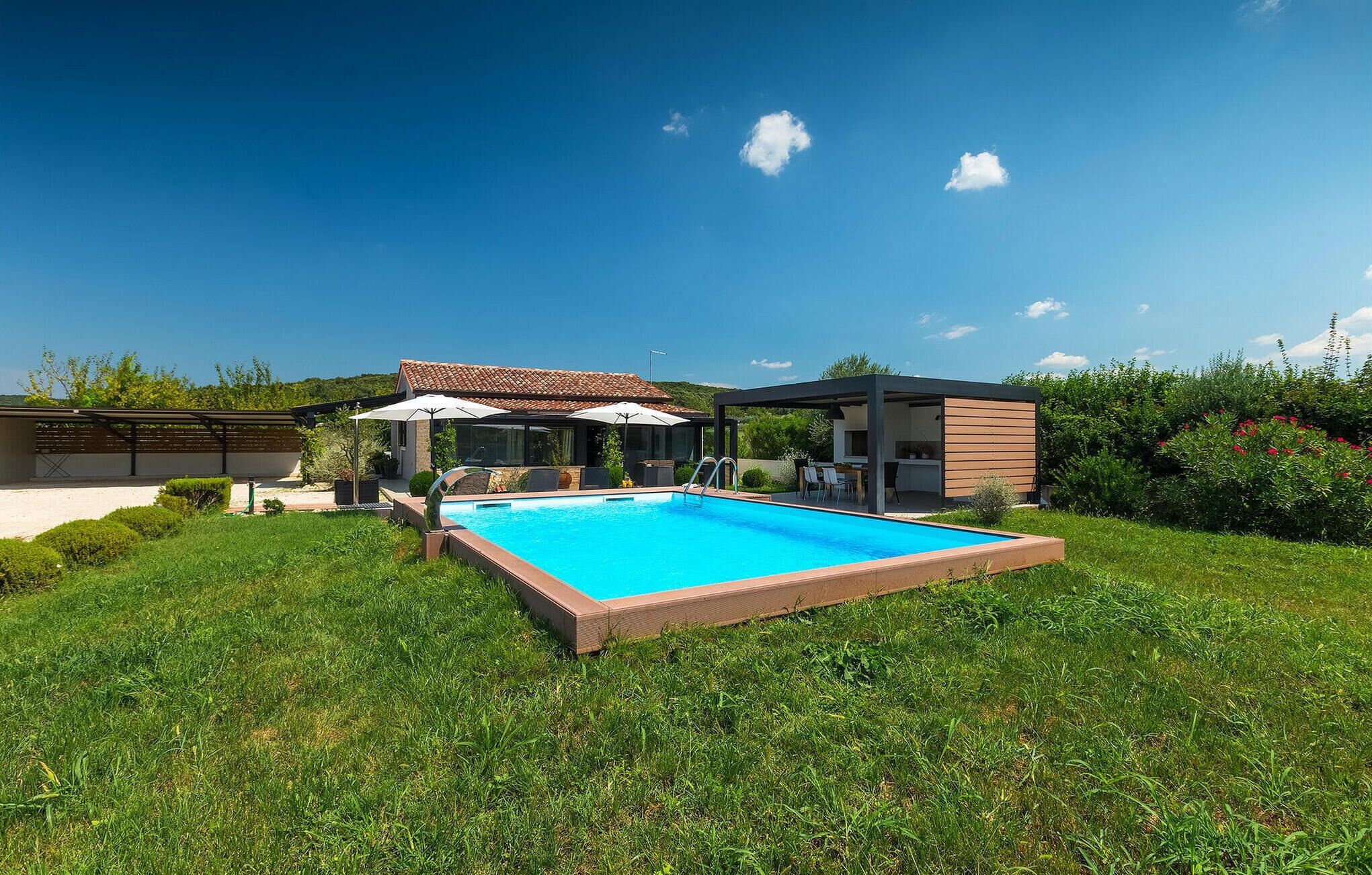 A modern outdoor swimming pool with a clear blue sky, surrounded by lush green grass and a small building with a tiled roof.