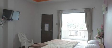 Un dormitorio con una cama de madera, una silla blanca, un televisor montado en la pared y una ventana con cortinas.